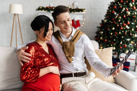 Photo for Happy man with ultrasound scan hugging pregnant asian woman in elegant clothes near blurred Christmas tree - Royalty Free Image
