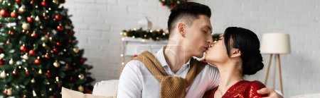 Foto de Joven hombre besos morena asiático esposa cerca de navidad árbol en borrosa fondo, bandera - Imagen libre de derechos
