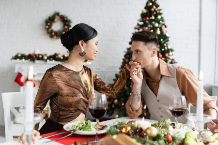 tätowierter Mann küsst die Hand einer asiatischen Frau im eleganten Outfit beim romantischen Abendessen zu Weihnachten zu Hause