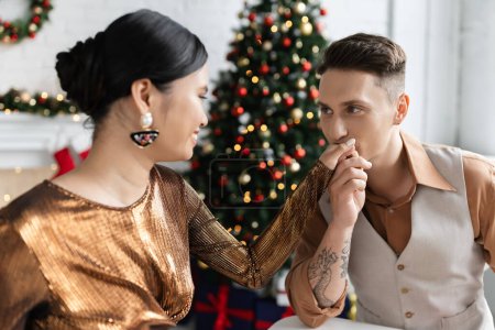 Photo for Tattooed man kissing hand of smiling asian wife in elegant shiny blouse - Royalty Free Image