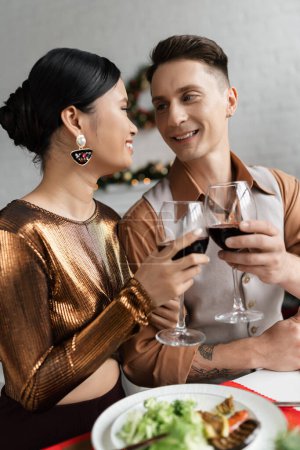 alegre pareja multiétnica en traje elegante mirándose unos a otros y tintinear copas de vino durante la cena romántica