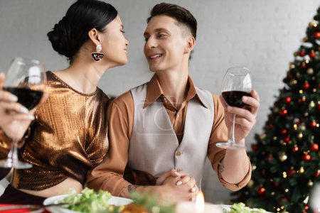 Foto de Pareja interracial en ropa festiva tomados de la mano y sonriendo el uno al otro mientras sostiene copas de vino - Imagen libre de derechos
