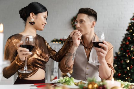 Foto de Hombre tatuado besos mano de elegante asiático esposa mientras celebrando la Navidad en casa - Imagen libre de derechos