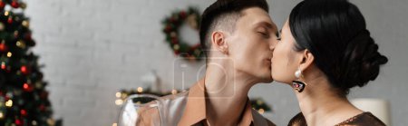 Foto de Joven pareja multiétnica besándose con los ojos cerrados mientras se celebra la Navidad en casa, pancarta - Imagen libre de derechos