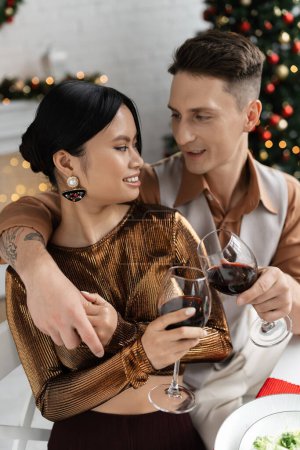 Foto de Elegante pareja multiétnica tintineo copas de vino durante la cena romántica festiva en casa - Imagen libre de derechos