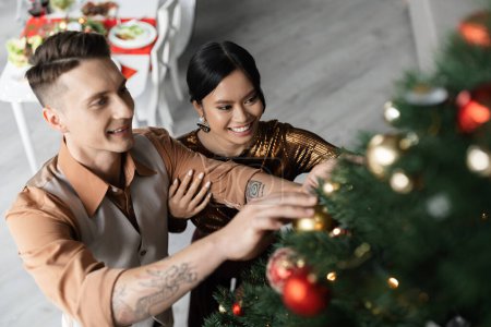 Téléchargez les photos : Vue grand angle de sourire interracial couple décoration arbre de Noël avec des boules brillantes - en image libre de droit
