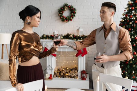 tattooed man giving glass of wine to happy asian wife during Christmas celebration 