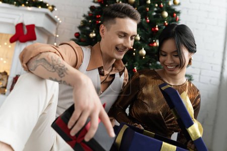 Foto de Feliz pareja interracial abriendo regalo de Navidad mientras está sentado bajo abeto decorado - Imagen libre de derechos