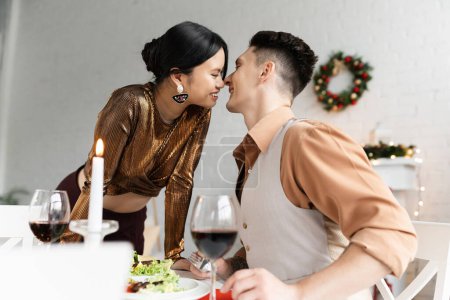 Foto de Feliz pareja interracial sonriendo cerca de la cena festiva en la mesa de comedor durante la celebración de Navidad - Imagen libre de derechos