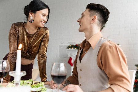 cheerful interracial couple looking at each other near festive meal and glasses of wine during Christmas 