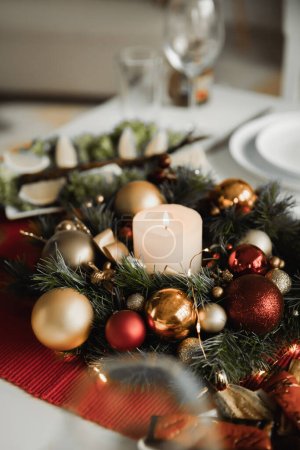 Corona navideña festiva con vela encendida y adornos brillantes en la mesa de comedor servida 