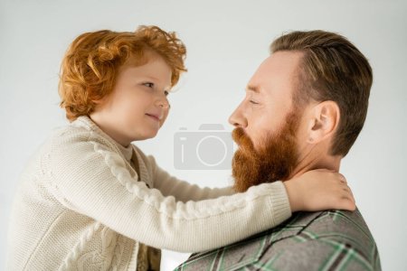 Téléchargez les photos : Souriant enfant roux étreignant père barbu en veste isolé sur gris - en image libre de droit