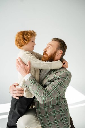Excited man in jacket hugging smiling redhead son on grey background