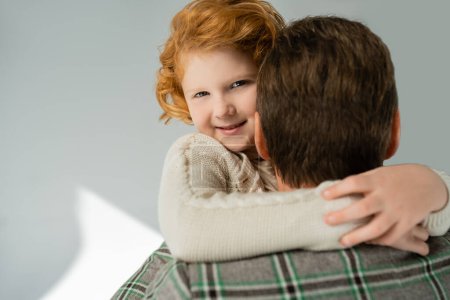 Cheerful red haired boy in knitted jumper hugging blurred dad on grey background