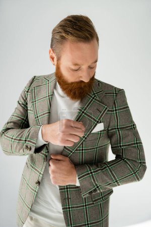 Elegante hombre barbudo mirando solapa de chaqueta sobre fondo gris