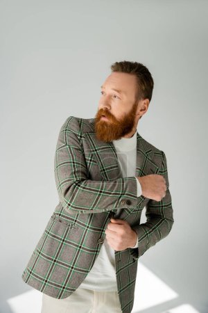 Fashionable bearded man adjusting jacket on grey background with sunlight 