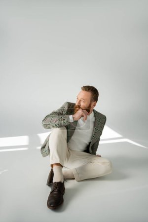 Foto de Hombre barbudo con chaqueta a cuadros mirando hacia otro lado mientras está sentado sobre fondo gris con luz solar - Imagen libre de derechos