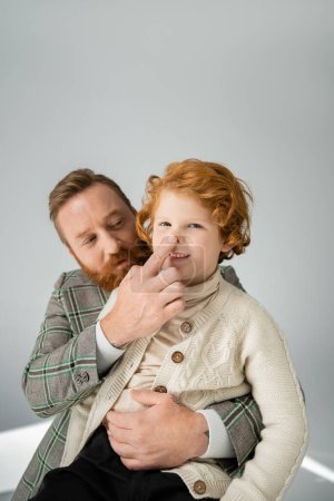Bearded man in jacket touching nose of red haired son on grey background
