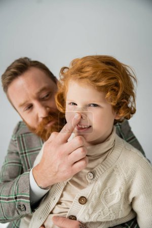 Blurred dad in jacket touching nose of red haired son isolated on grey 