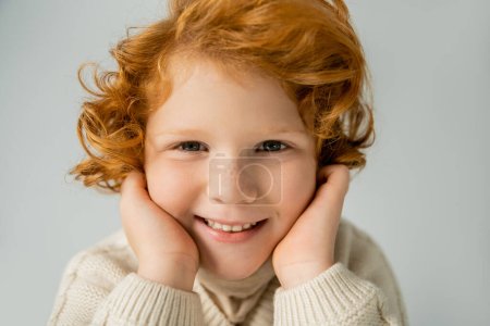 Cheerful red haired kid in beige jumper touching cheeks isolated on grey 