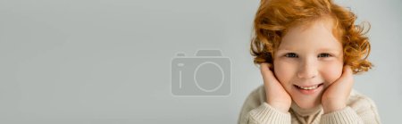 Portrait of positive red haired boy in jumper looking at camera isolated on grey, banner 