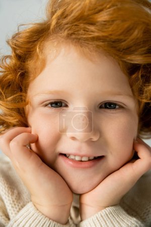 Close up view of smiling redhead kid in knitted jumper touching cheeks isolated on grey 