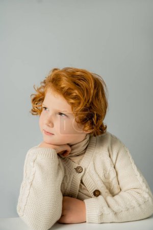 Freckled and redhead boy in knitted jumper looking away near table isolated on grey 