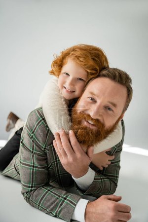 Photo for Red haired boy hugging father while lying on grey background with sunlight - Royalty Free Image