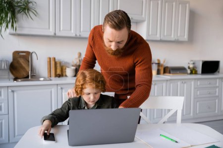 bärtiger Mann erklärt seinem rothaarigen Sohn beim Hausaufgabenmachen in der Nähe des Laptops etwas