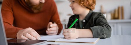 Téléchargez les photos : Rousse garçon tenant stylo près de notebook tout en faisant des devoirs avec papa, bannière - en image libre de droit