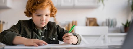 Photo for Redhead kid holding pen near blurred notebook while doing homework in kitchen, banner - Royalty Free Image