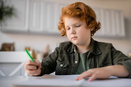 pensive redhead boy holding pen while doing homework near blurred notebook