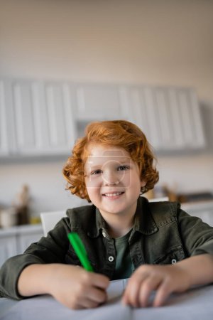 happy redhead kid with pen looking at camera while doing homework on blurred foreground