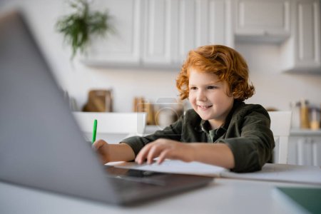 fröhlicher rothaariger Junge macht Hausaufgaben in der Nähe eines verschwommenen Laptops in der Küche