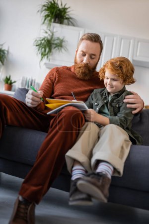 Photo for Redhead boy writing in copybook near bearded dad on couch at home - Royalty Free Image