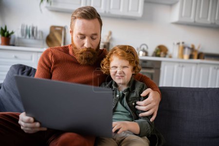 bored boy frowning near laptop and redhead dad on couch at home