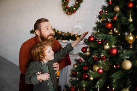 bearded man holding son reaching baubles on decorated Christmas tree