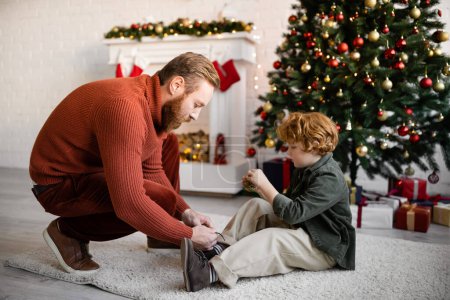 Foto de Vista lateral del niño pelirrojo sentado en el suelo cerca de papá atando sus cordones y el árbol de Navidad con chimenea decorada - Imagen libre de derechos