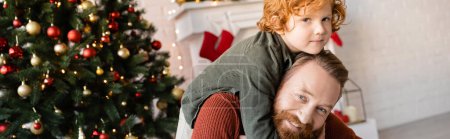 happy bearded man piggybacking redhead son near blurred Christmas tree, banner