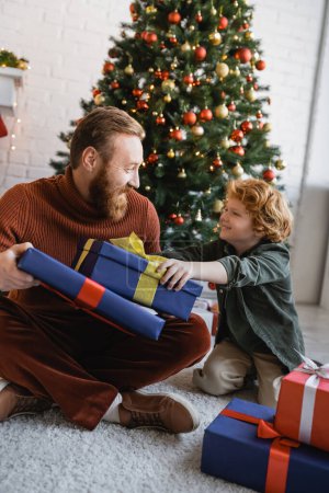 glücklicher rothaariger Vater und Sohn schauen einander in der Nähe von Geschenkschachteln und Weihnachtsbaum zu Hause an