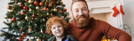 happy redhead boy with bearded father looking at camera near decorated Christmas tree, banner