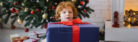 amazed redhead kid looking at camera near big gift box and Christmas tree, banner