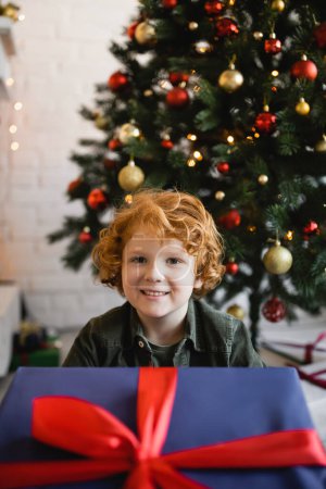 zufriedener Junge mit roten Haaren blickt in die Kamera in der Nähe großer Geschenkschachtel und Weihnachtsbaum auf verschwommenem Hintergrund