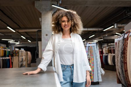 Photo for Cheerful and curly saleswoman standing near desk in textile shop - Royalty Free Image