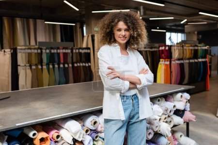 Photo for Cheerful and curly saleswoman standing with crossed arms near rack with different fabric rolls in textile shop - Royalty Free Image