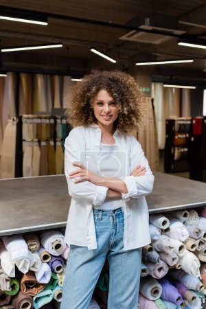 vendedora feliz de pie con brazos cruzados cerca de rack con diferentes rollos de tela en la tienda textil 