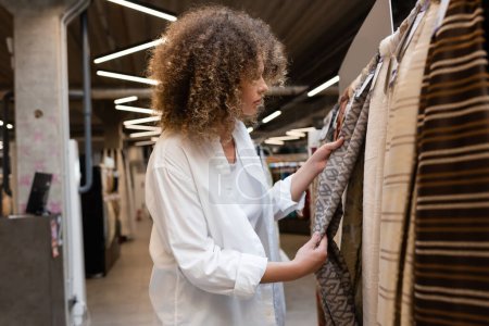 Téléchargez les photos : Vue latérale de la vendeuse bouclée vérifiant le matériau du tissu dans l'atelier textile - en image libre de droit