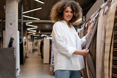 happy and curly saleswoman checking patterned fabric in textile shop 