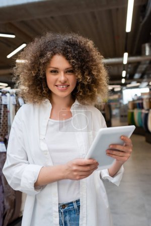 glückliche Verkäuferin lächelt, während sie im Textilgeschäft ein digitales Tablet in der Hand hält 