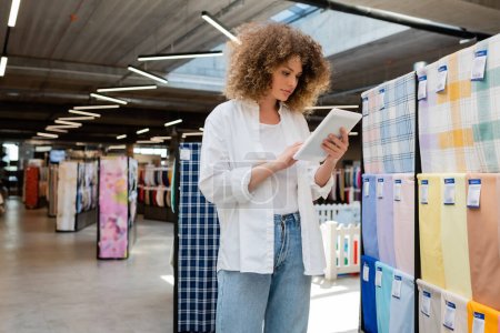 joven vendedora utilizando tableta digital cerca de tela estampada en la tienda textil 